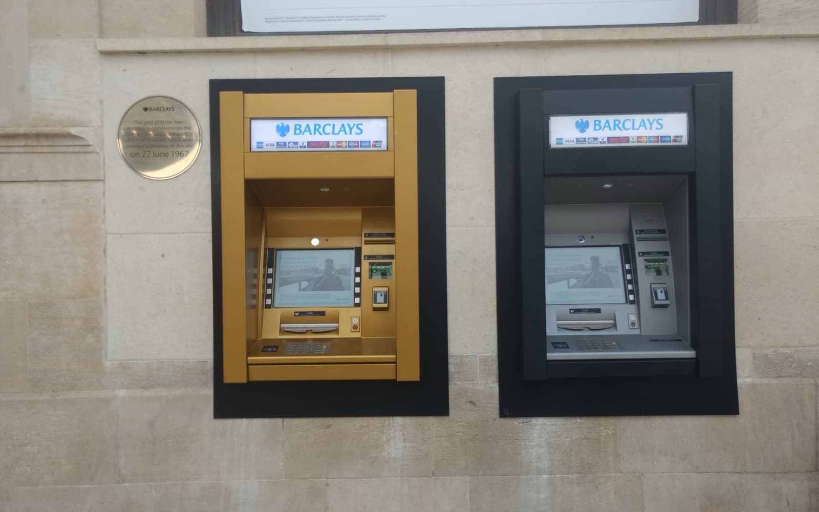 Two cash machines set into a stone wall, one painted gold, with a small commemorative plaque beside them