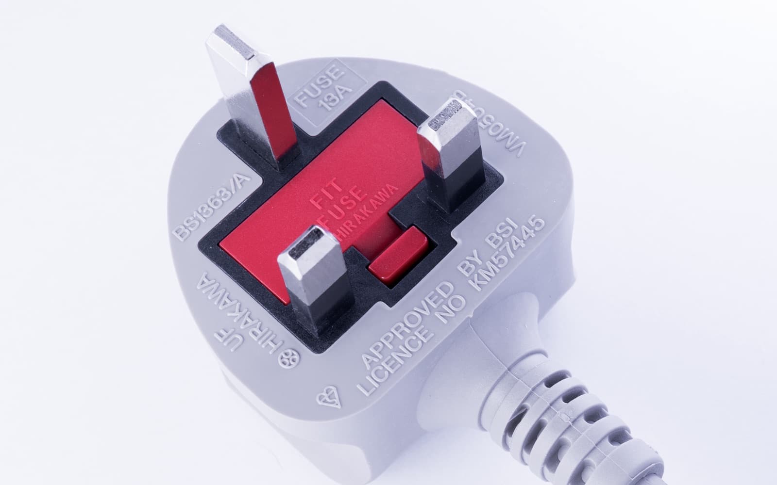 Close-up of a grey moulded British three-pin plug with flat rectangular pins and a red fuse cover marked Fit Fuse, with Approved by BSI printed on it
