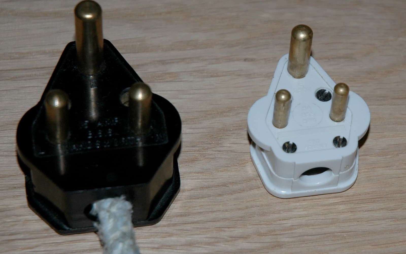 Two old British round-pin plugs on a wooden table, a large black 15 amp plug with a fabric-covered cord and a smaller white 5 amp plug