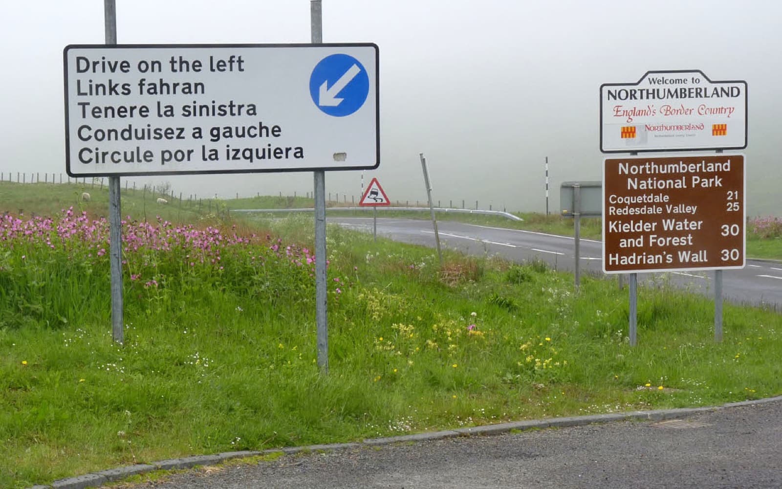Roadside sign reading Drive on the left in English, German, Italian, French and Spanish, with a blue keep-left arrow, beside a Welcome to Northumberland sign on a misty moorland road