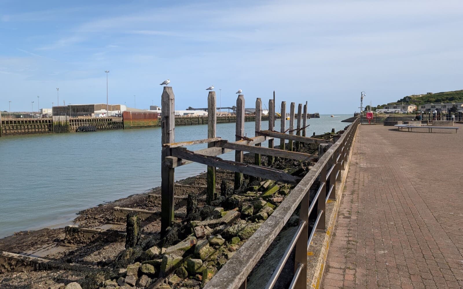A harbour wall with old wooden posts and a river mouth leading out to sea under a blue sky