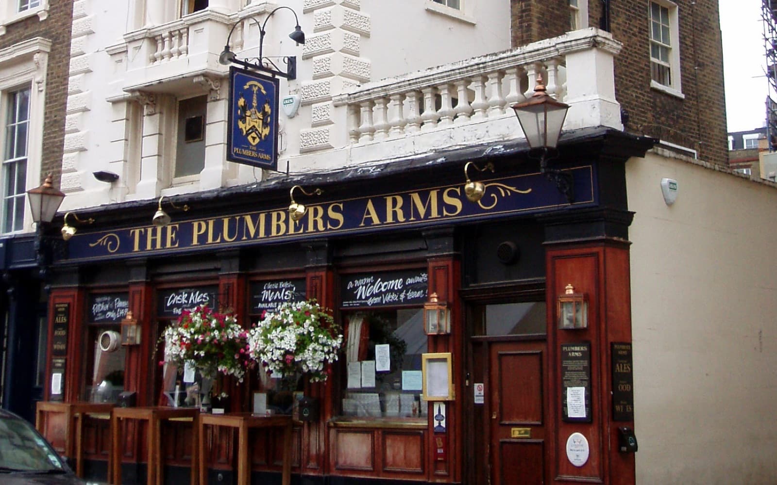 The front of a traditional London pub with a dark painted frontage and a sign reading The Plumbers Arms