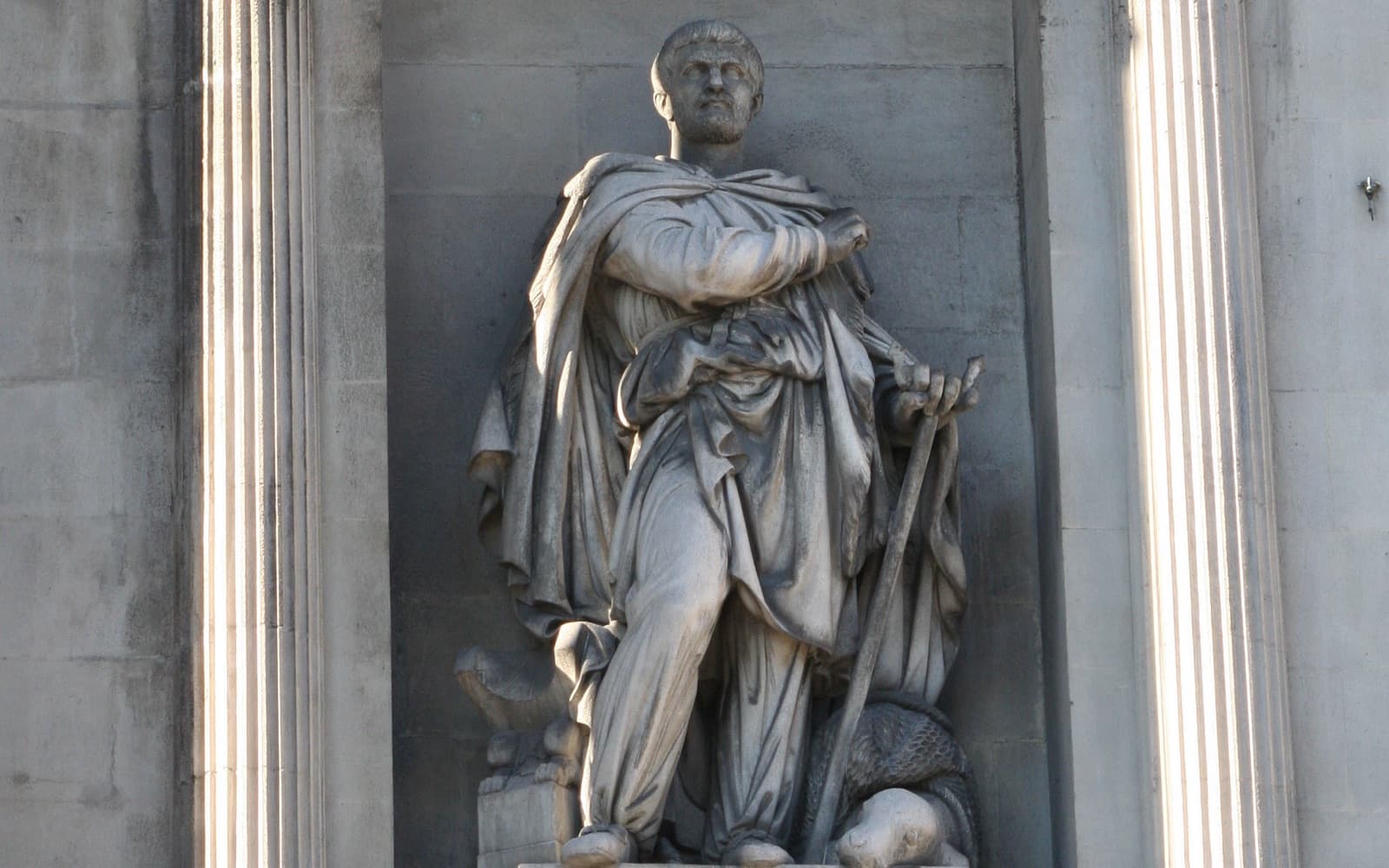 Stone statue of a bearded man in a heavy cloak, holding a staff, standing in a niche between two fluted columns