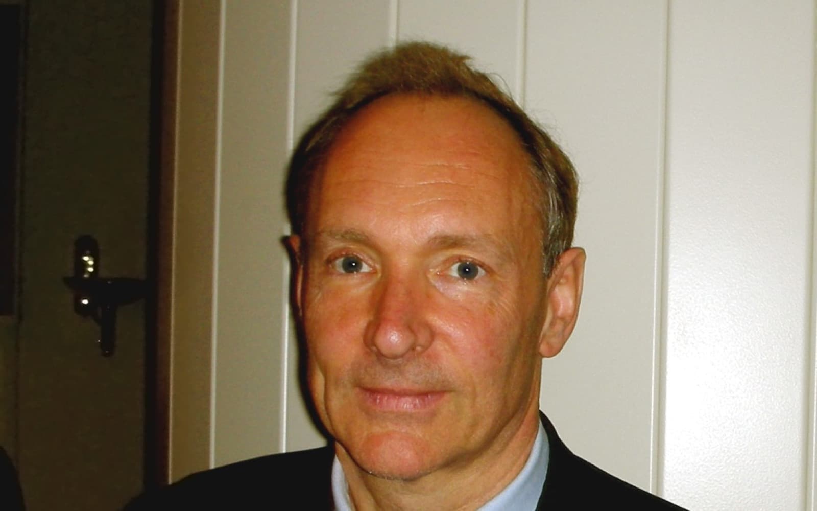 Head-and-shoulders photo of Tim Berners-Lee in a dark jacket and light blue shirt, standing in front of a white panelled wall