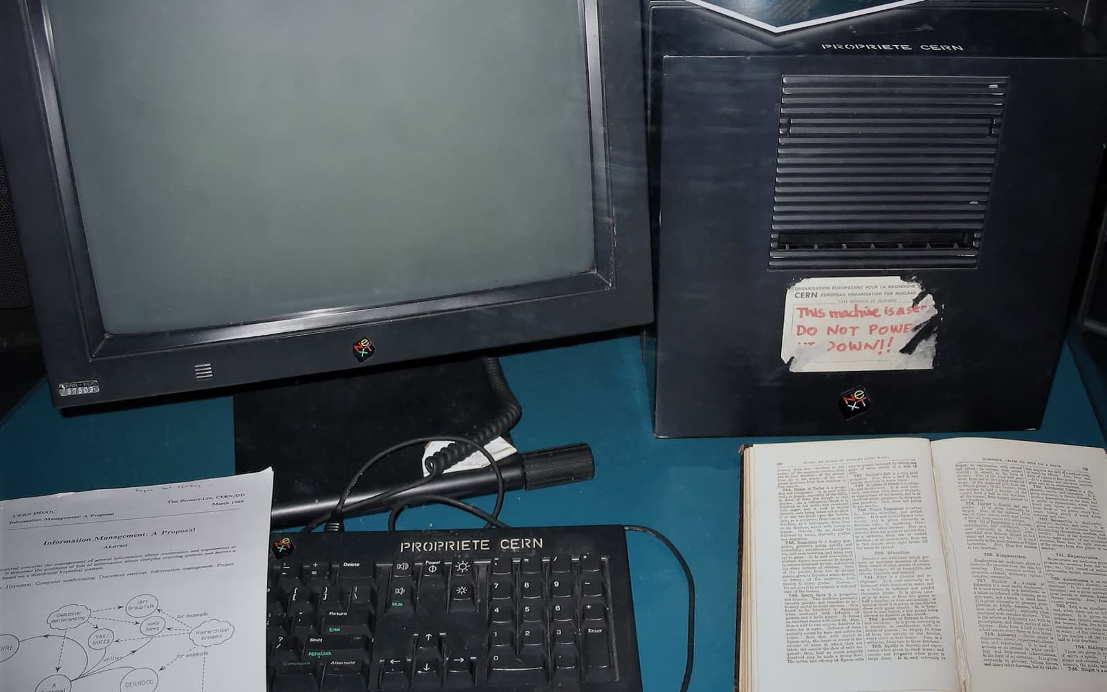 A black NeXT computer and monitor in a glass case, with a torn label on the computer reading "This machine is a server. DO NOT POWER IT DOWN!!", next to a printed proposal and an open old book