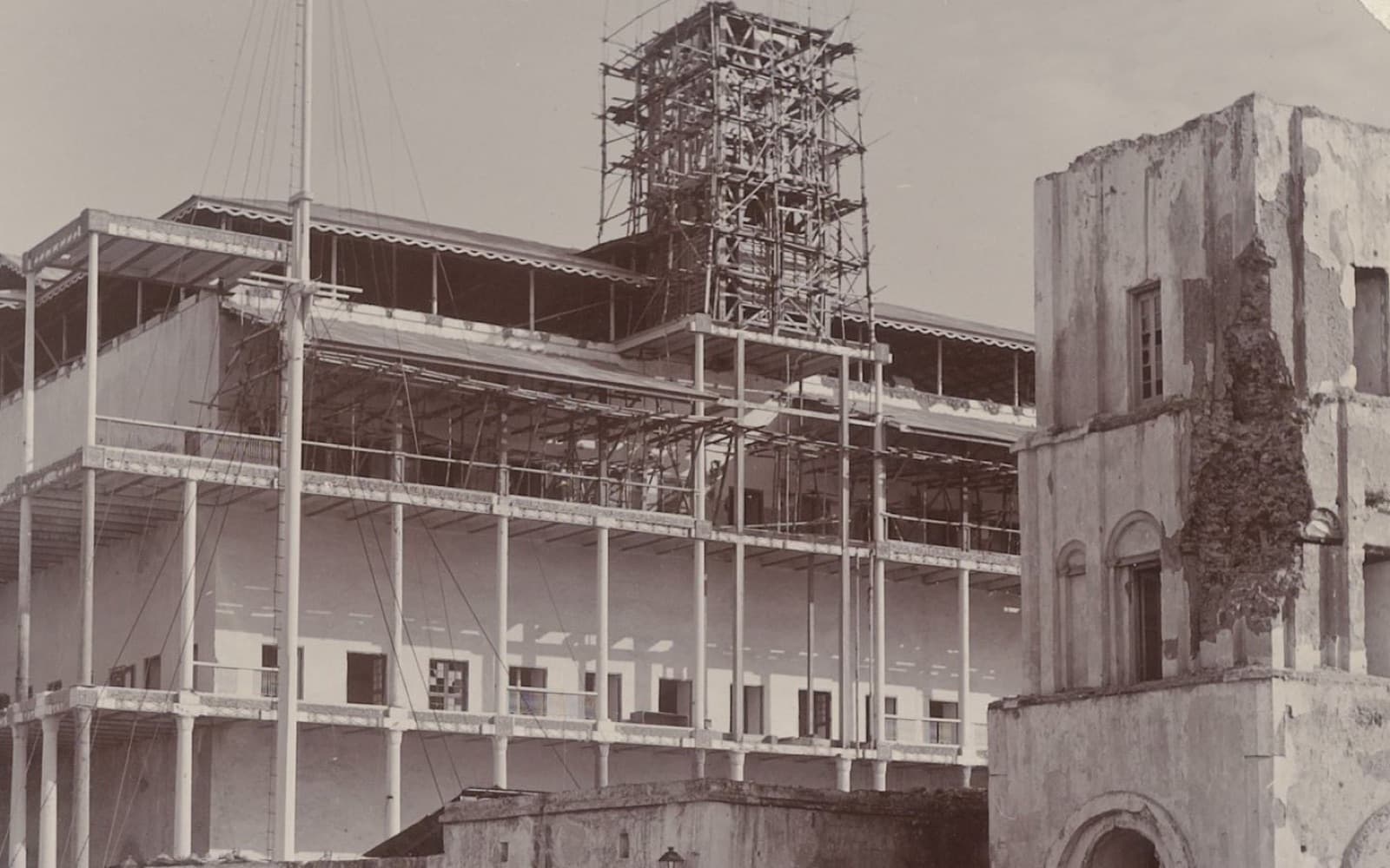 Old photograph of a large waterfront building under scaffolding next to a damaged stone building